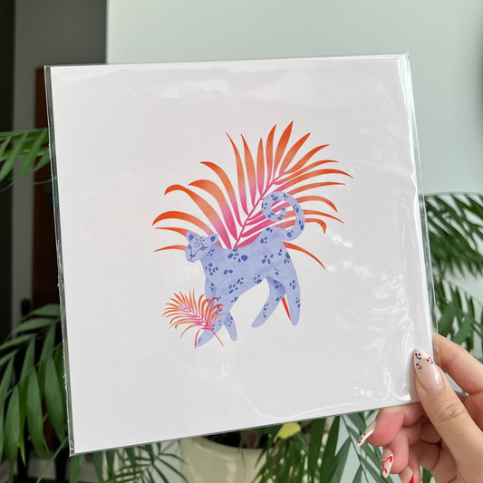 Hand holding a white square with a colorful design of a leopard and plants against a light background.