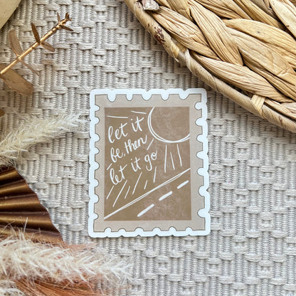Decorative sticker shaped like a stamp with text 'let it be, let it be, let it go' on a textured surface with dried plants.