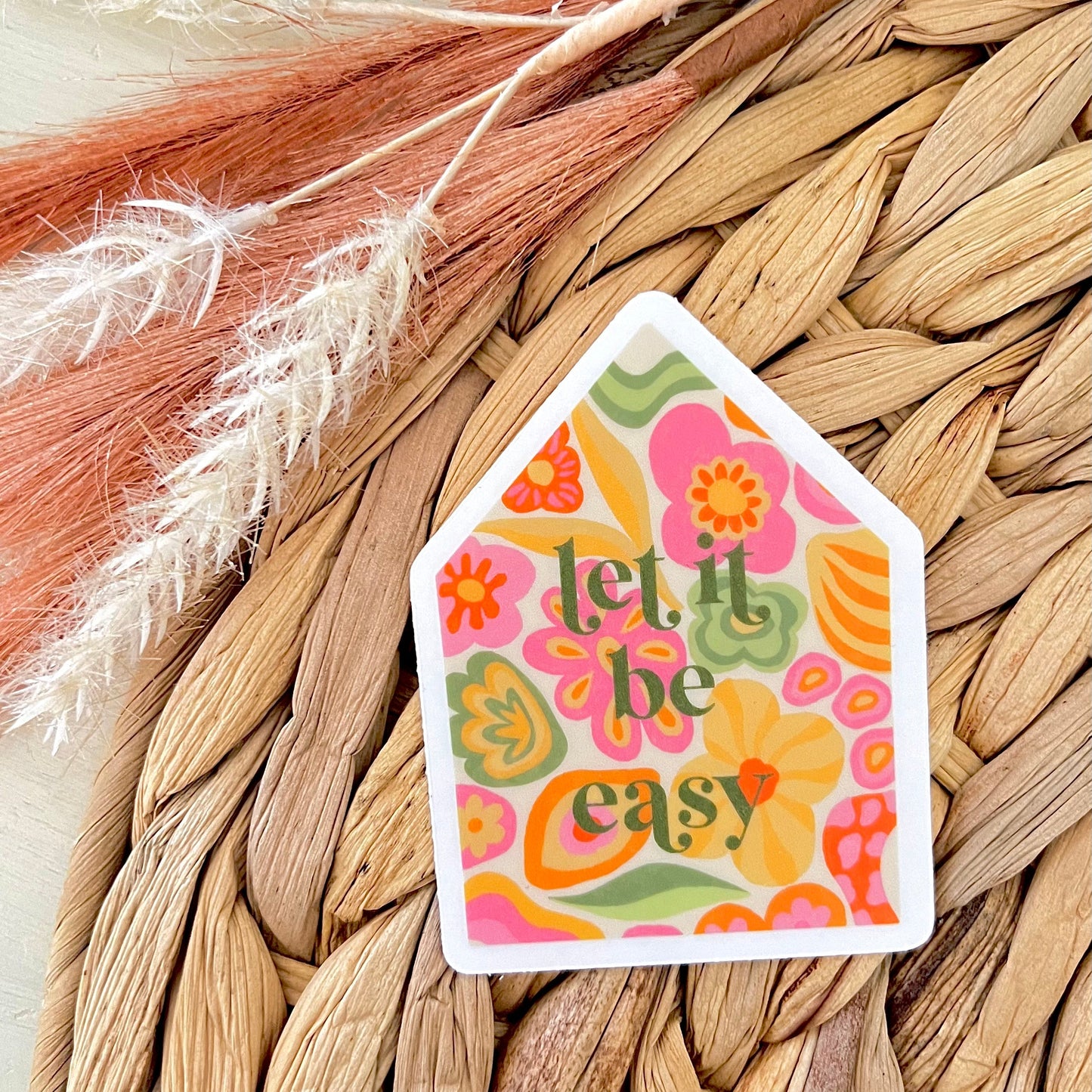 Decorative house-shaped sticker with floral design and 'let it be easy' text on a woven surface.