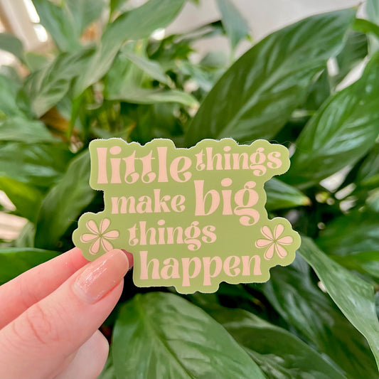 Hand holding a green sticker with 'little things make big things happen' text against a leafy background