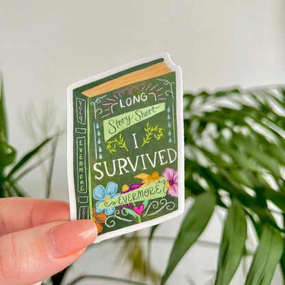 Hand holding a book-themed sticker with decorative text and illustrations against a plant background