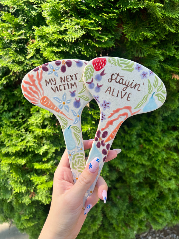 Two colorful ceramic garden stakes with decorative designs and text held against a green bush background.