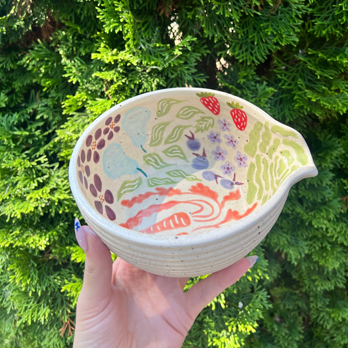 Hand holding a ceramic bowl with colorful patterns against a green foliage background