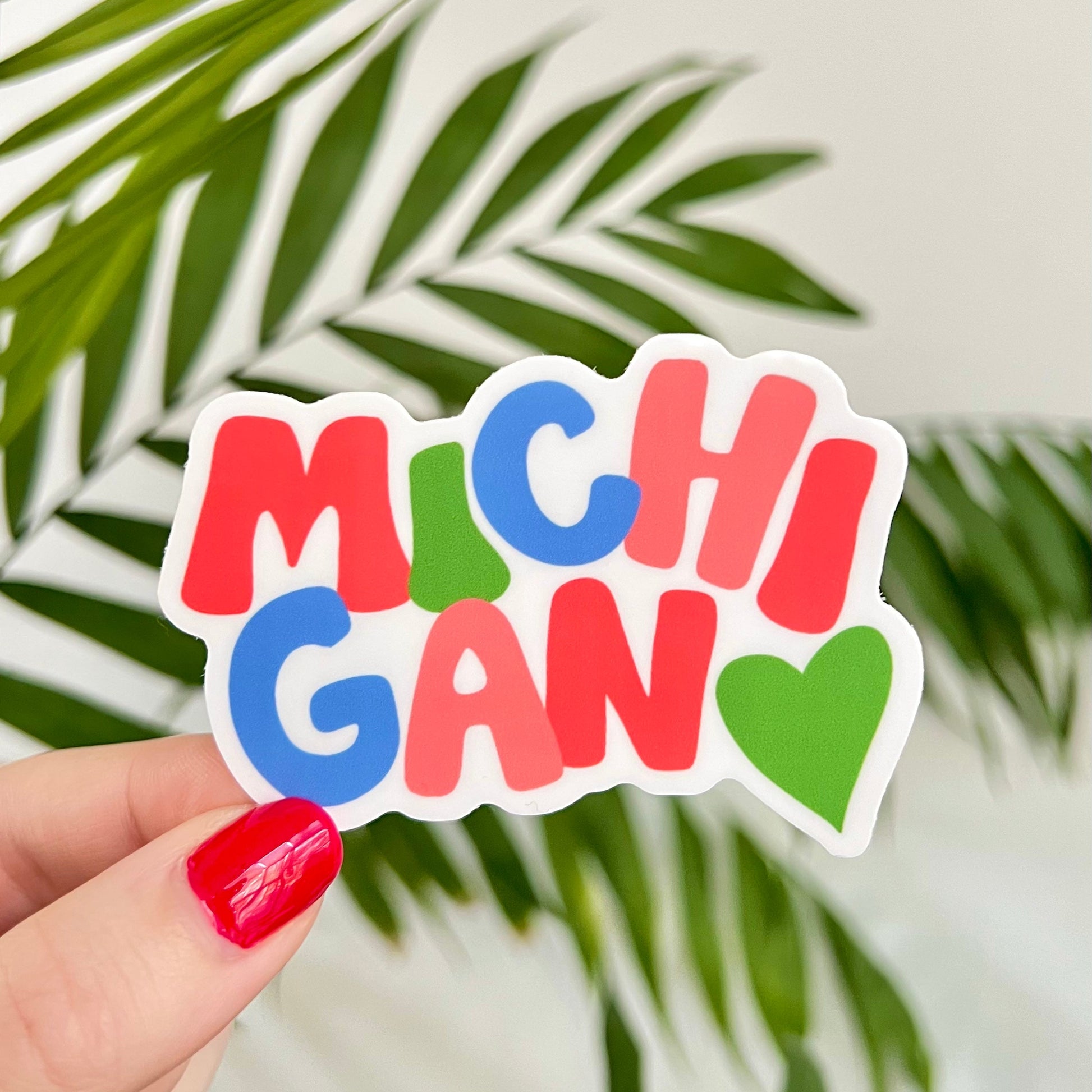 Colorful 'MICHIGAN' sticker held by a hand with red nails against a leafy background.