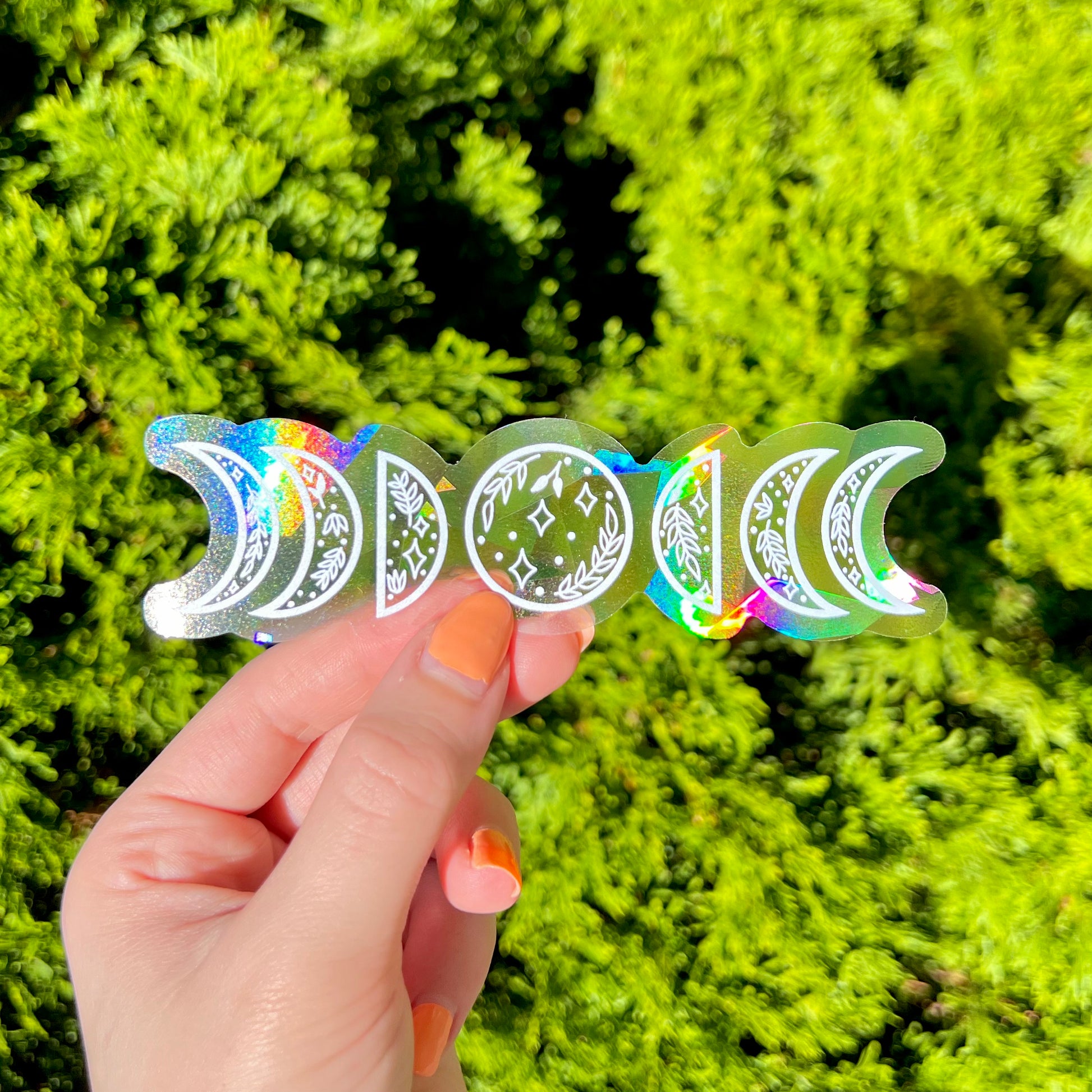 Hand holding holographic moon phase suncatcher stickers against a green leafy background
