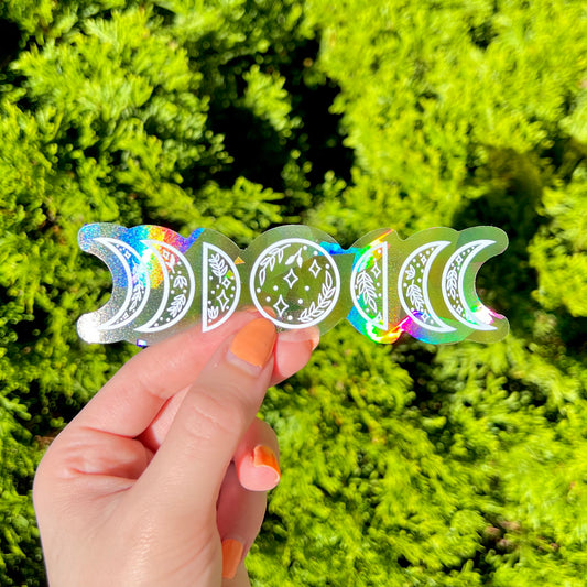Hand holding holographic moon phase suncatcher stickers against a green leafy background