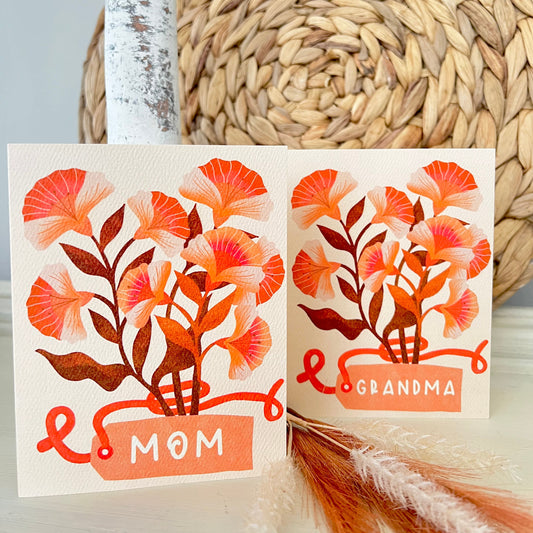 Two greeting cards with floral designs and text 'Mom' and 'Grandma' on a textured surface.