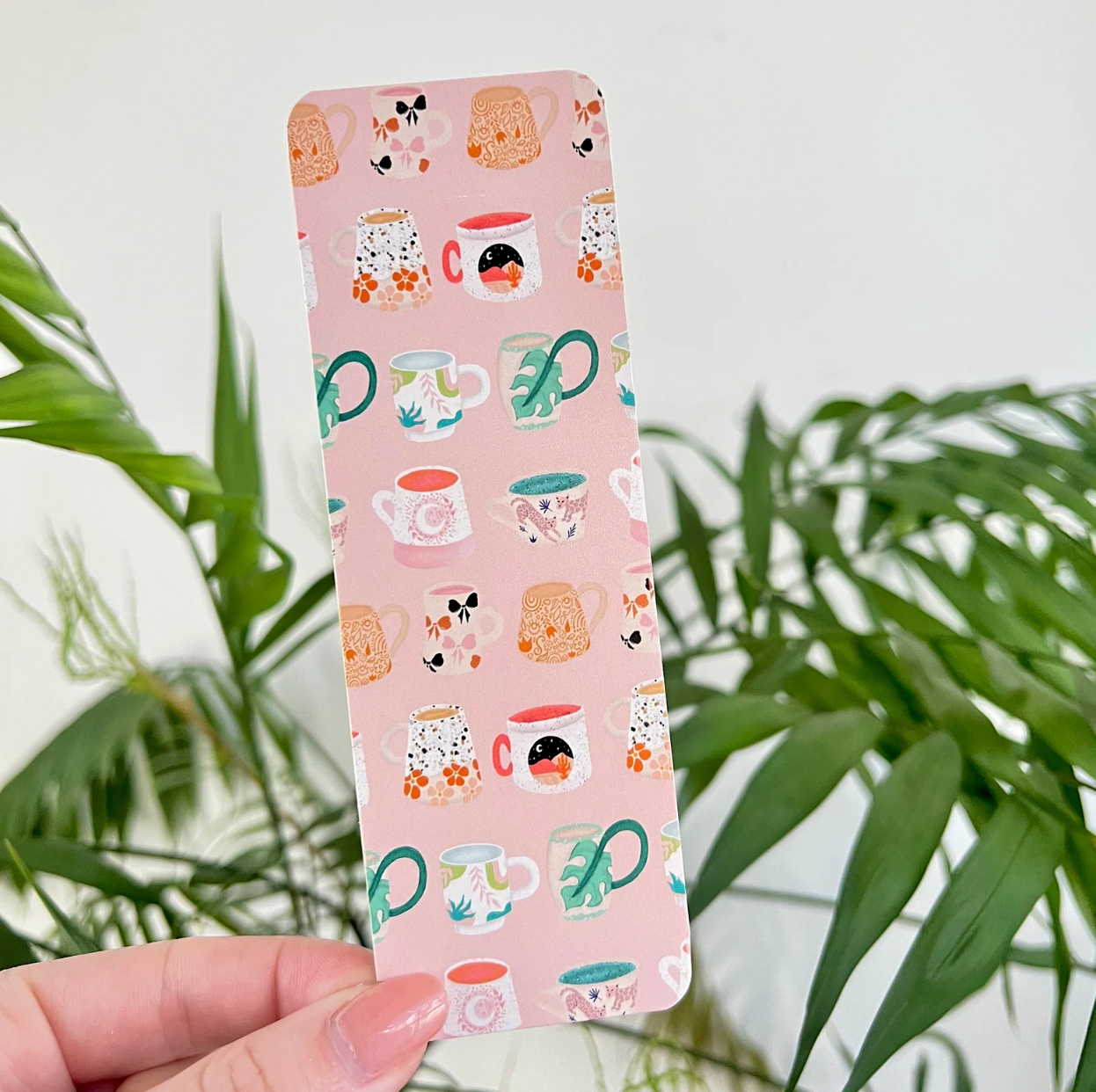 Pink bookmark with colorful mug pattern held by a hand against a green plant background