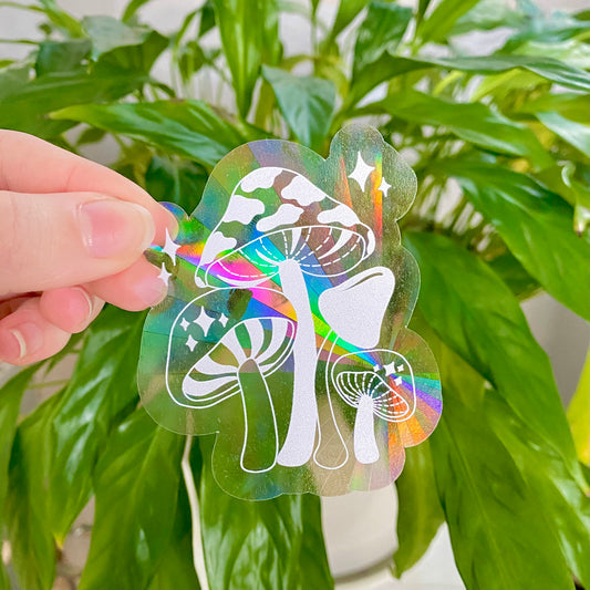 Hand holding a colorful mushroom-shaped suncatcher holographic sticker in front of green plants