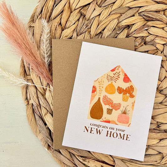 Greeting card with house illustration and 'congrats on your NEW HOME' text on a woven mat.