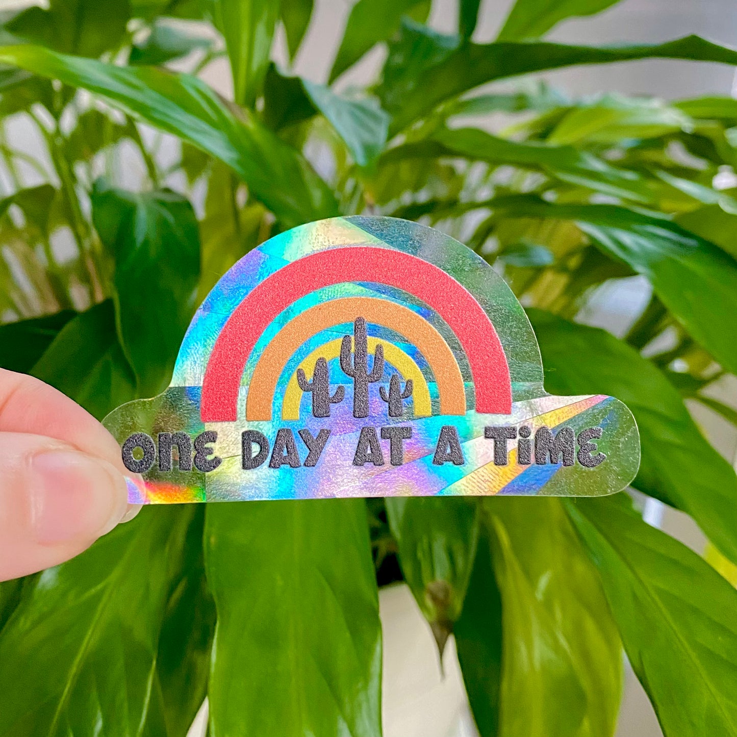 Colorful rainbow-shaped suncatcher sticker with 'One Day at a Time' text held in front of green leaves