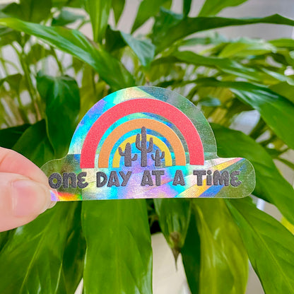 Colorful rainbow-shaped suncatcher sticker with 'One Day at a Time' text held in front of green leaves