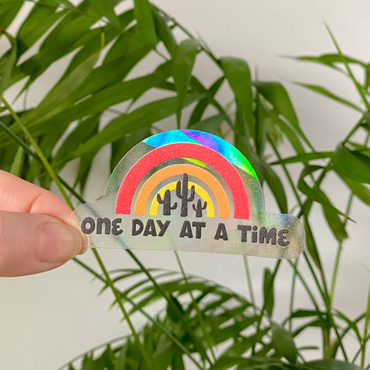 Holographic rainbow sticker with cacti and 'One Day at a Time' text held in front of green leaves.