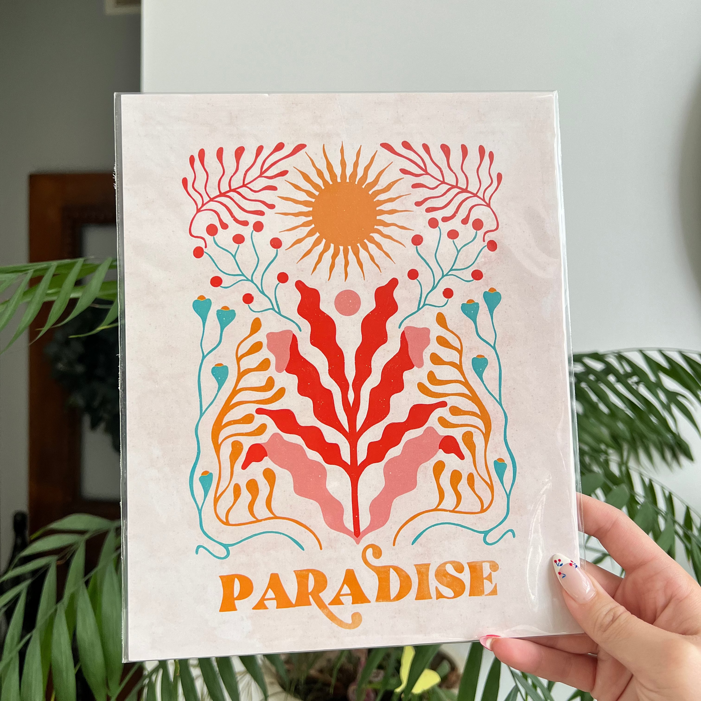 Handheld decorative print with floral design and 'Paradise' text, surrounded by greenery.
