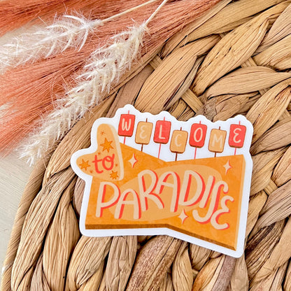 Sticker with 'Welcome to Paradise' text on a woven surface with decorative grasses.