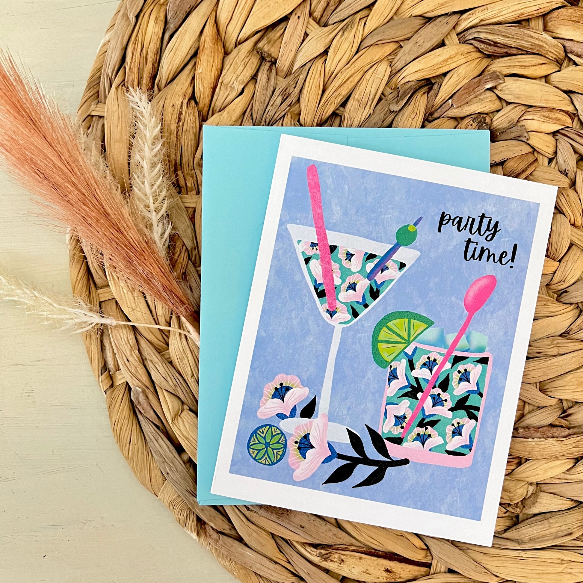 Greeting card with floral cocktail illustration and blue envelope on a woven mat