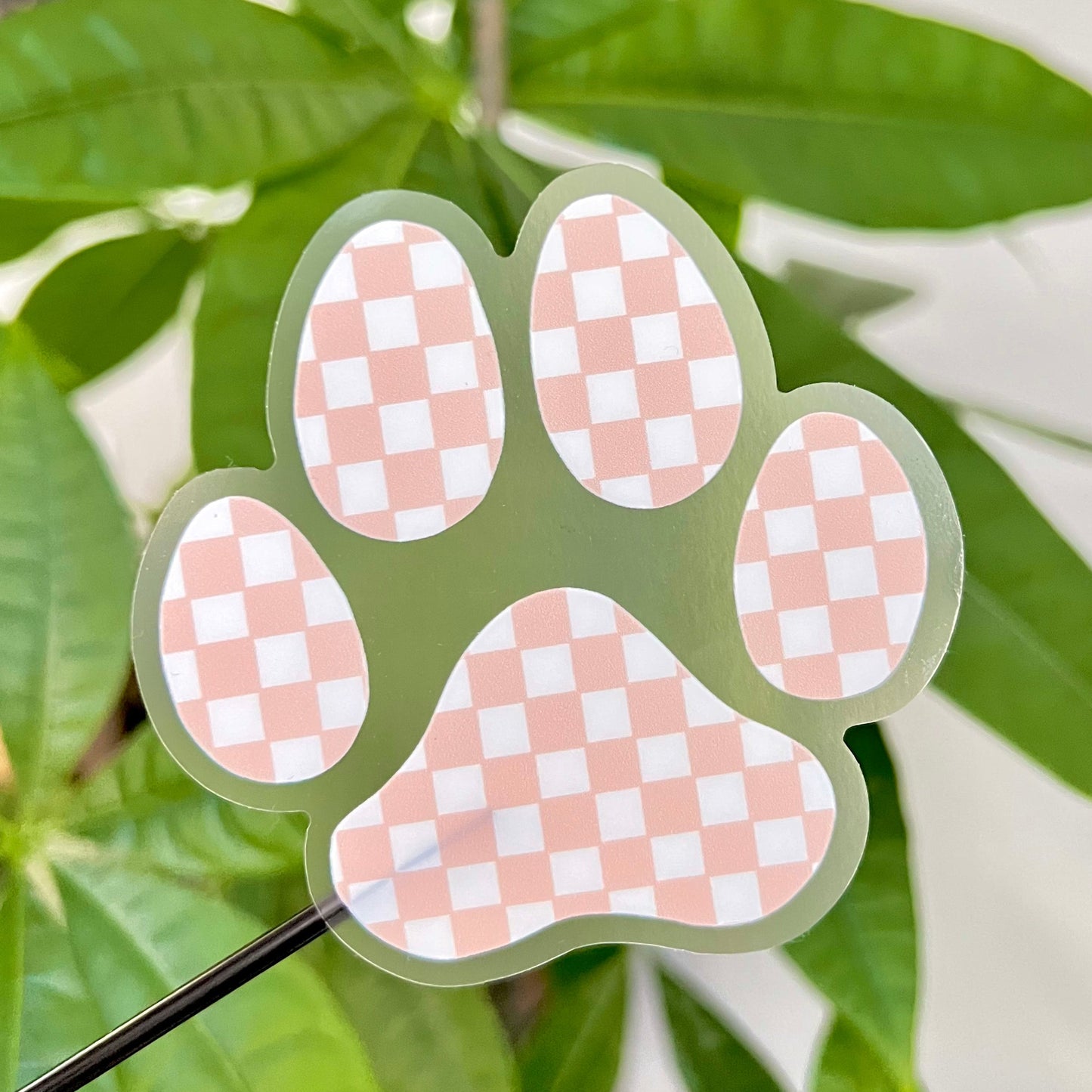 Checkered pink paw-shaped sticker on a green leafy background