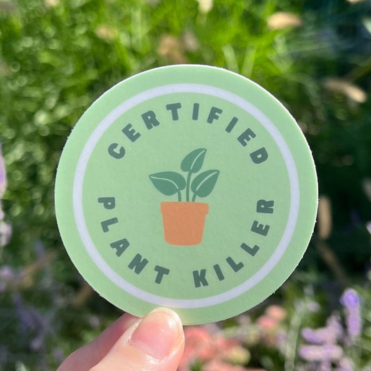 Green sticker with 'Certified Plant Killer' text held in front of a blurred natural background