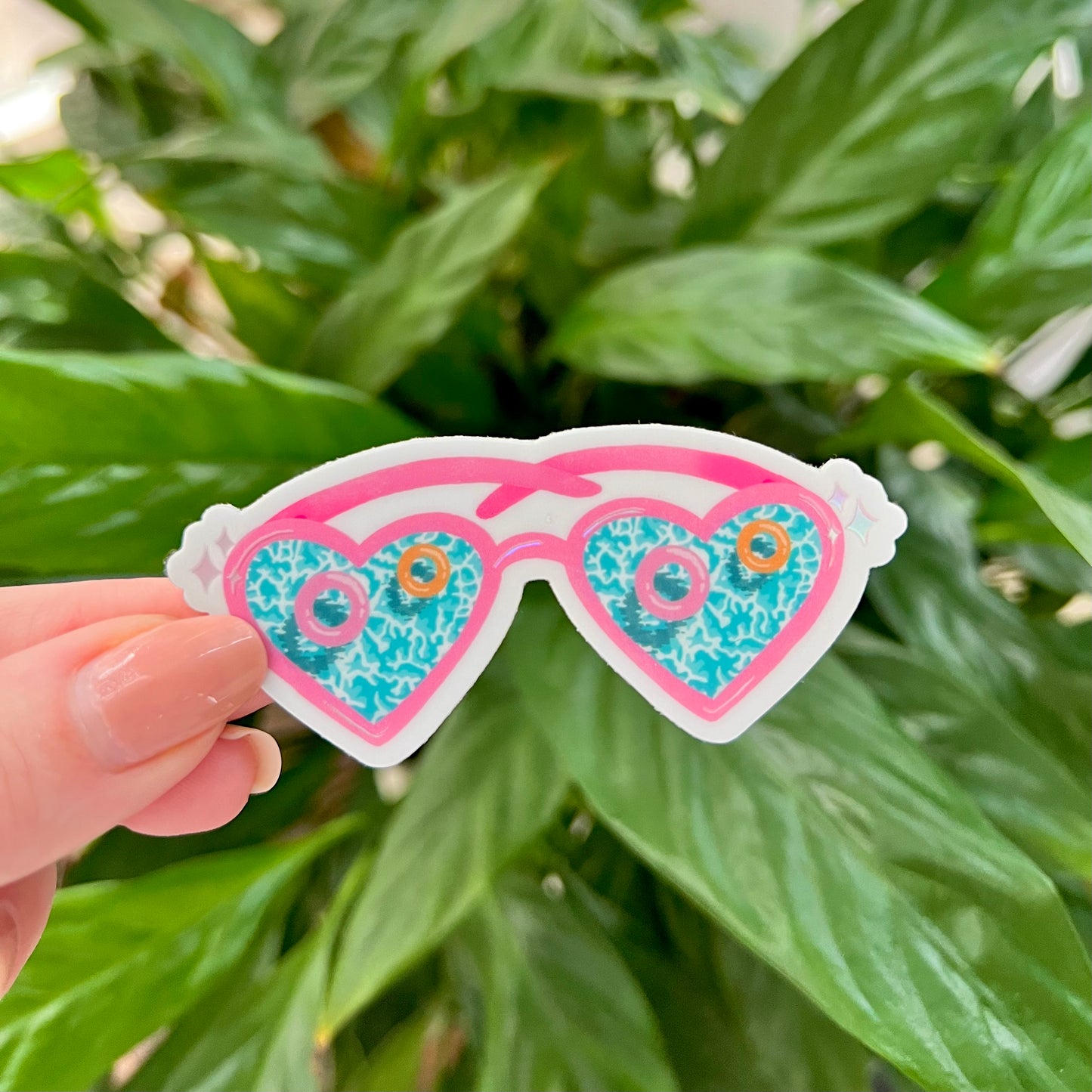 Heart-shaped sunglasses sticker held in front of green leaves