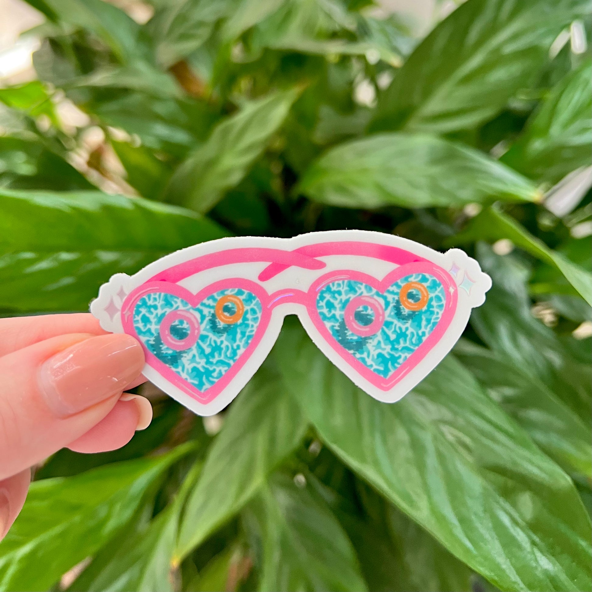 Heart-shaped sunglasses sticker held in front of green leaves