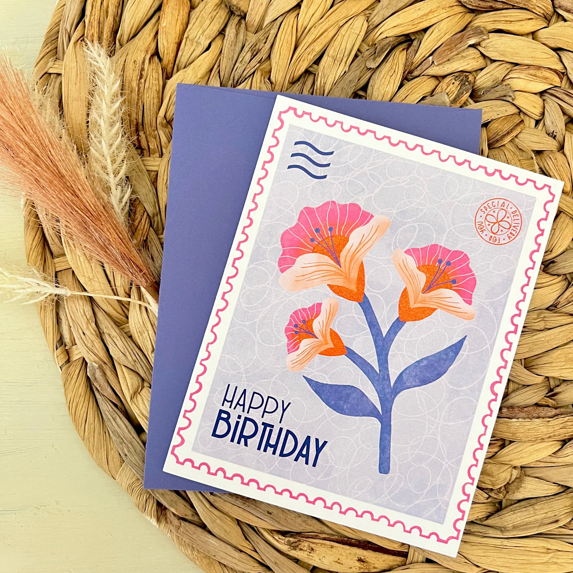 Birthday card with floral design and 'Happy Birthday' text and a purple envelope on a woven mat background