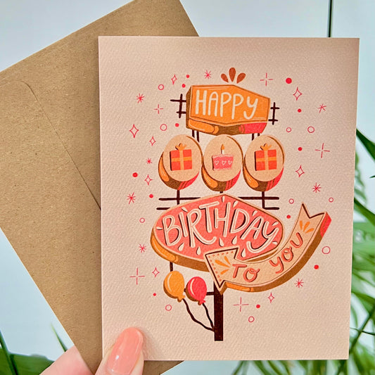 Hand holding a birthday card with illustration of retro sign that says "happy birthday to you"