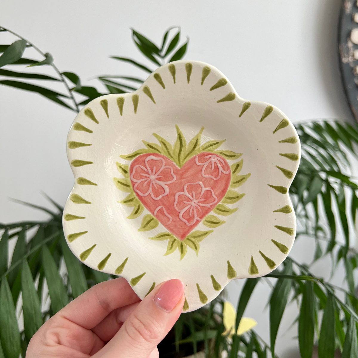 Hand holding a decorative plate with a heart design against a plant background