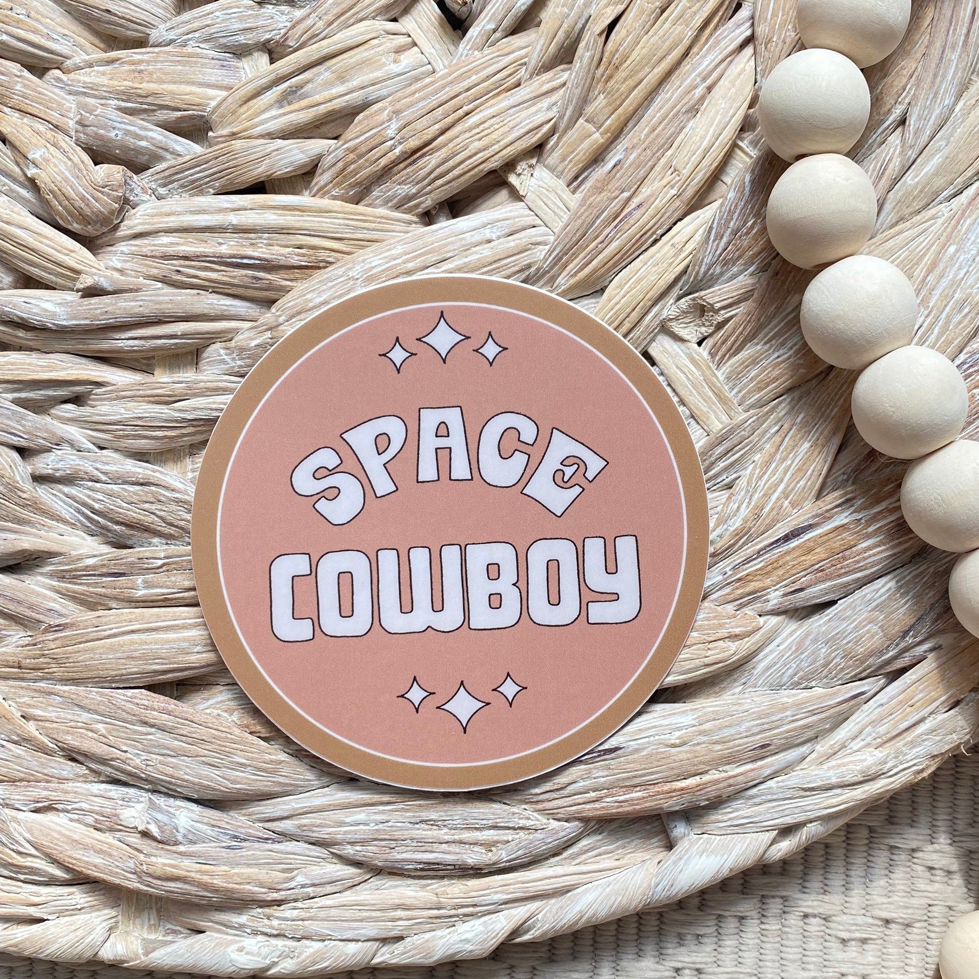 Round pink sticker with 'Space Cowboy' text on a woven mat with beads