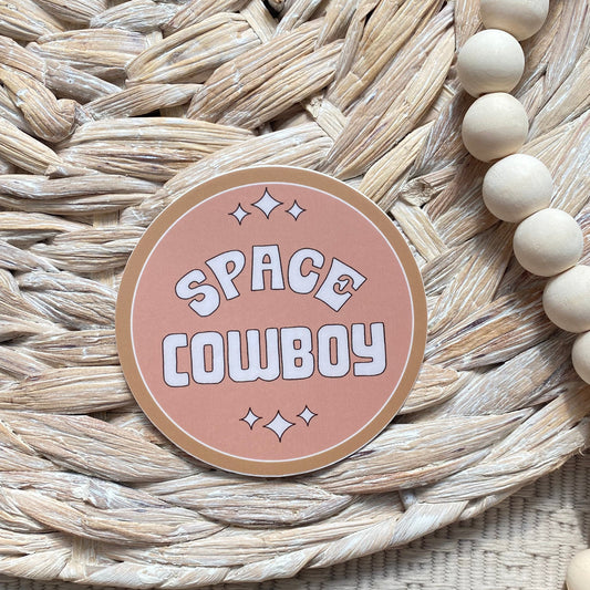 Round pink sticker with 'Space Cowboy' text on a woven mat with beads