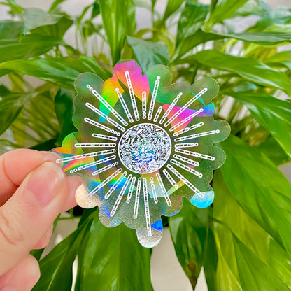 Holographic sun-shaped suncatcher sticker held in front of green leaves