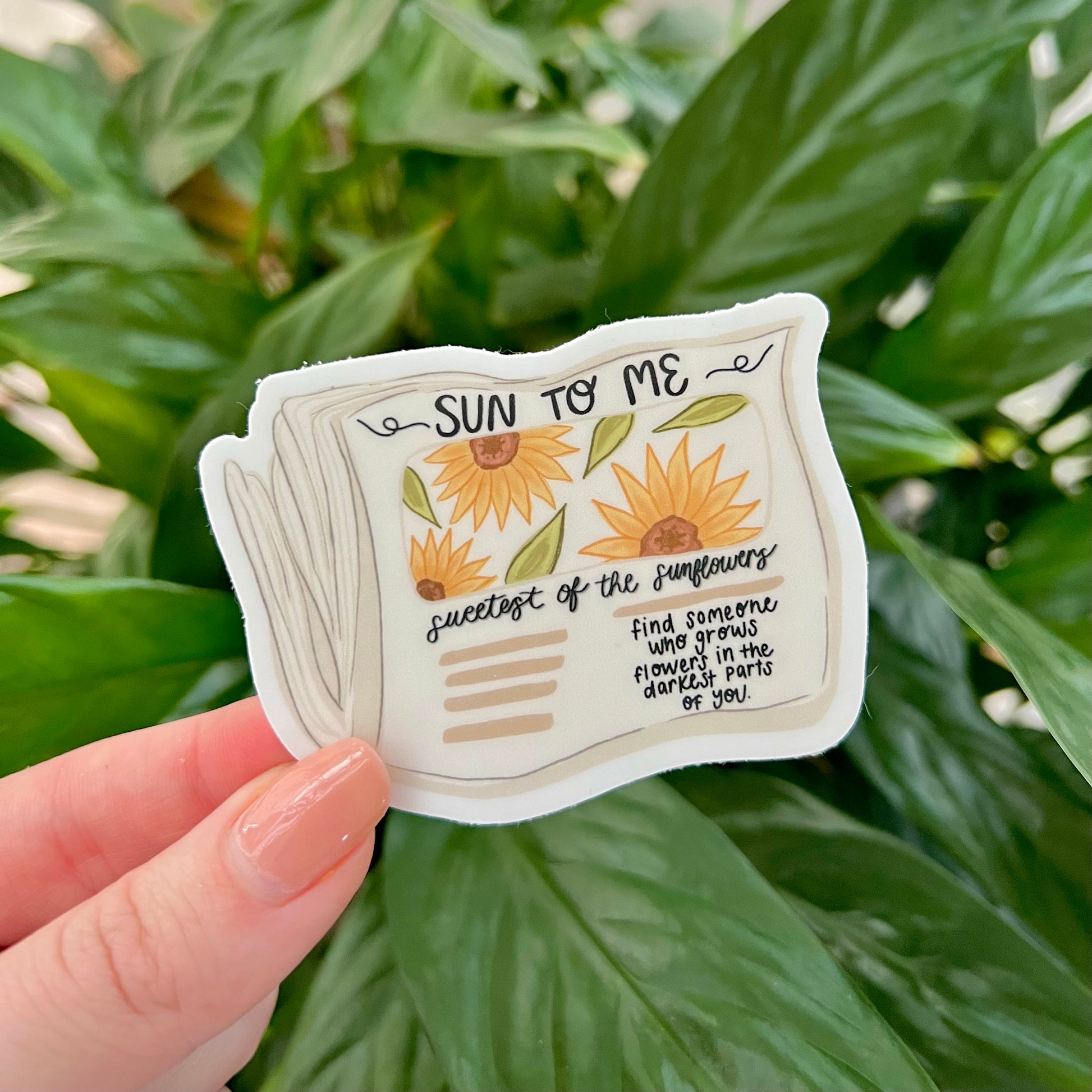Sticker of an illustration of newspaper saying "sun to me" and sunflowers in front of greenery