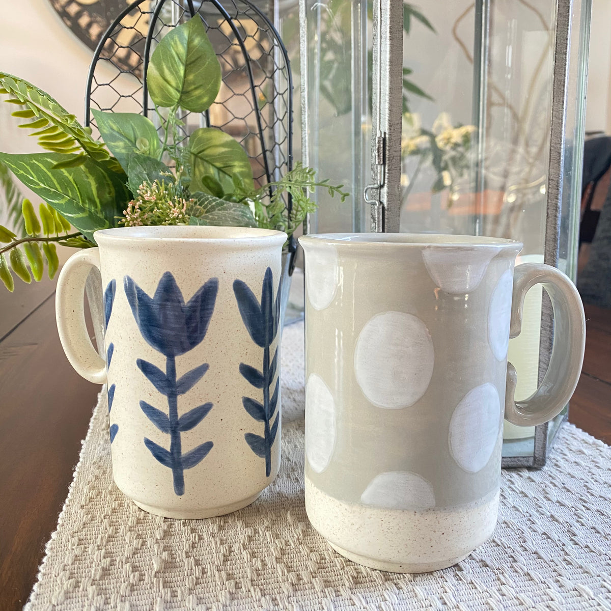 Two ceramic mugs with decorative patterns on a table with a plant in the background.