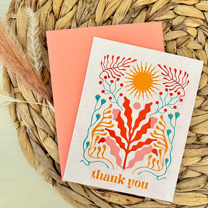 Greeting card with floral design and 'thank you' text and peach envelope on a woven mat