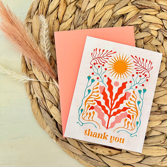 Greeting card with floral design and 'thank you' text and peach envelope on a woven mat