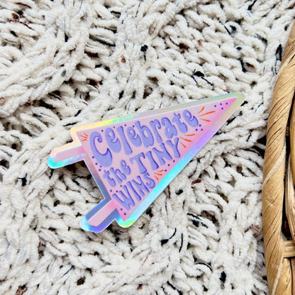 Holographic pennant flag sticker with "celebrate the tiny wins" text on it sitting on a textured surface