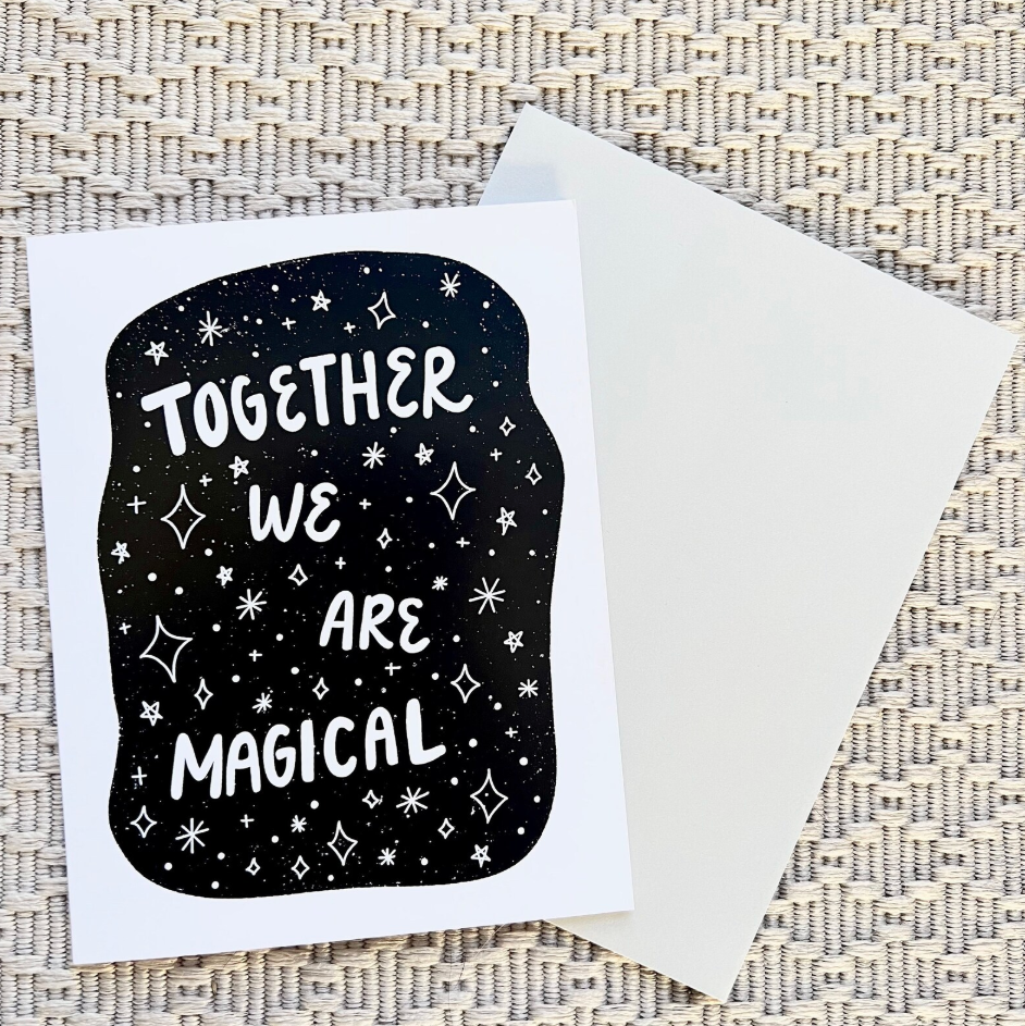Greeting card with 'Together We Are Magical' text on a black background, featuring stars, with a white envelope on a textured surface.