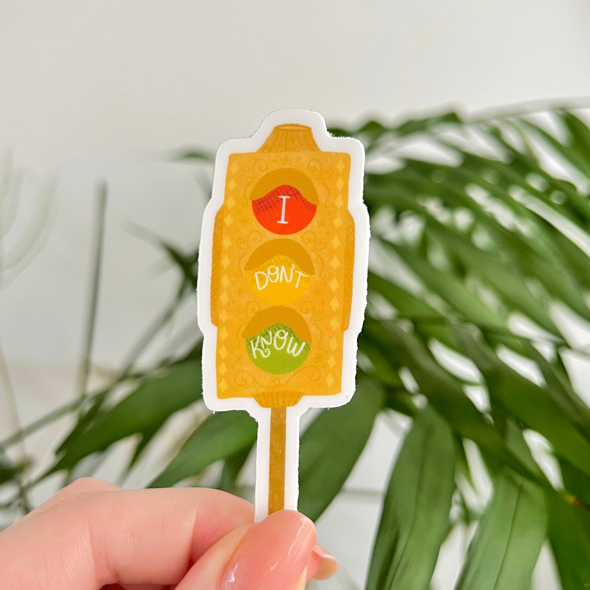 Hand holding a novelty stoplight with text 'I Don't Know' against a blurred green plant background