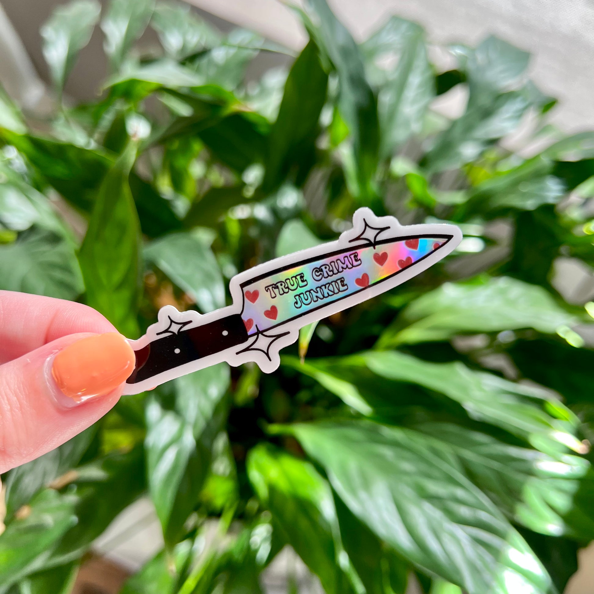 Knife shaped holographic sticker with "true crime junkie" written on it held in front of green leaves