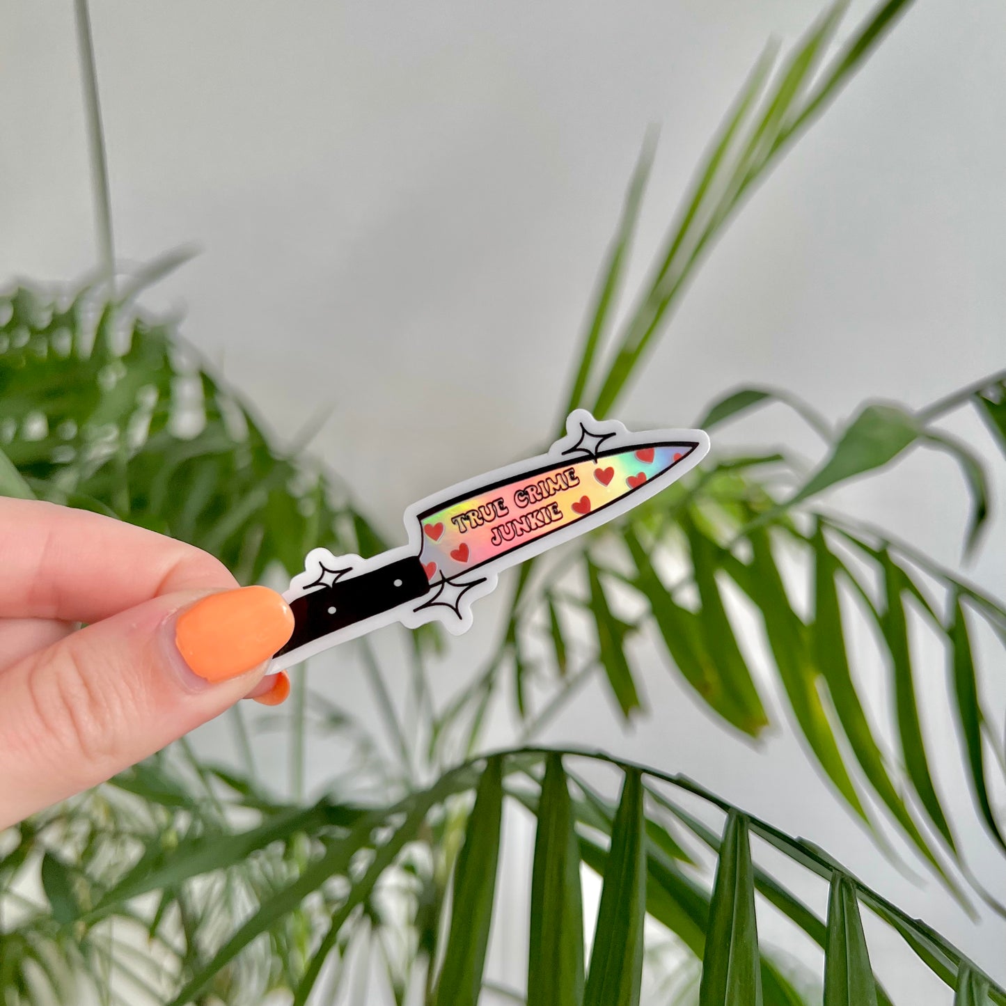 Hand holding a holographic knife-shaped sticker with text saying "true crime junkie" against a green leafy background