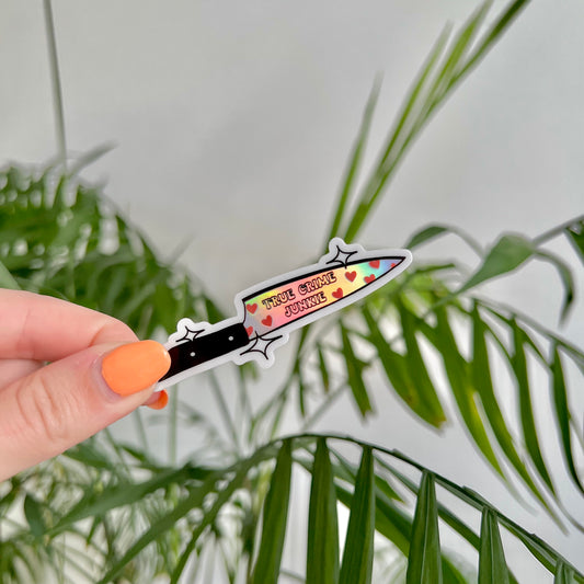 Hand holding a holographic knife-shaped sticker with text saying "true crime junkie" against a green leafy background