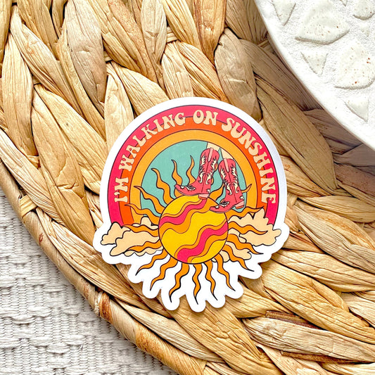 Sticker with a colorful design of a sun and text 'I'm Walking on Sunshine' on a woven mat.
