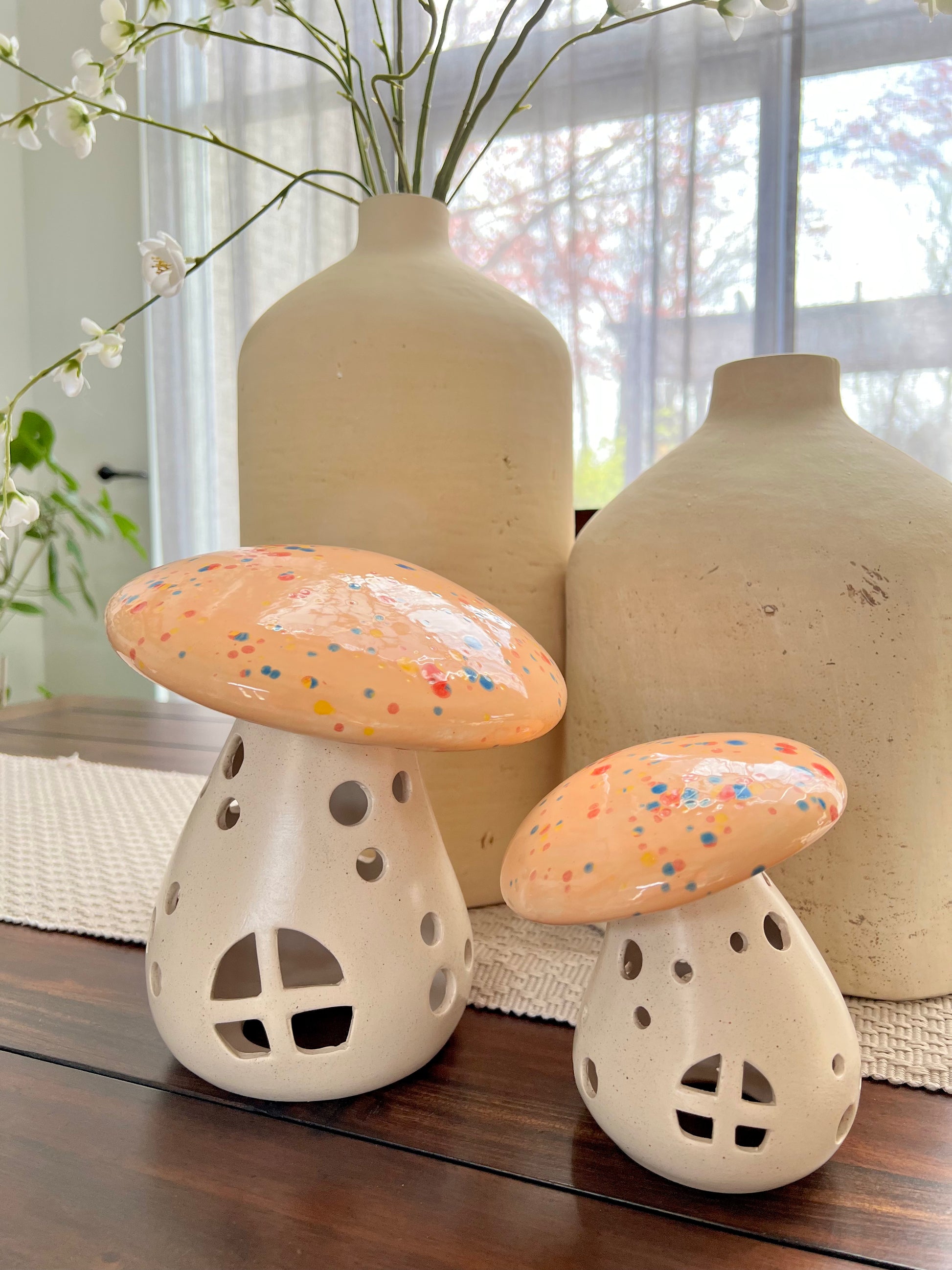 Two ceramic mushroom-shaped votives on a wooden surface with large beige vases in the background.