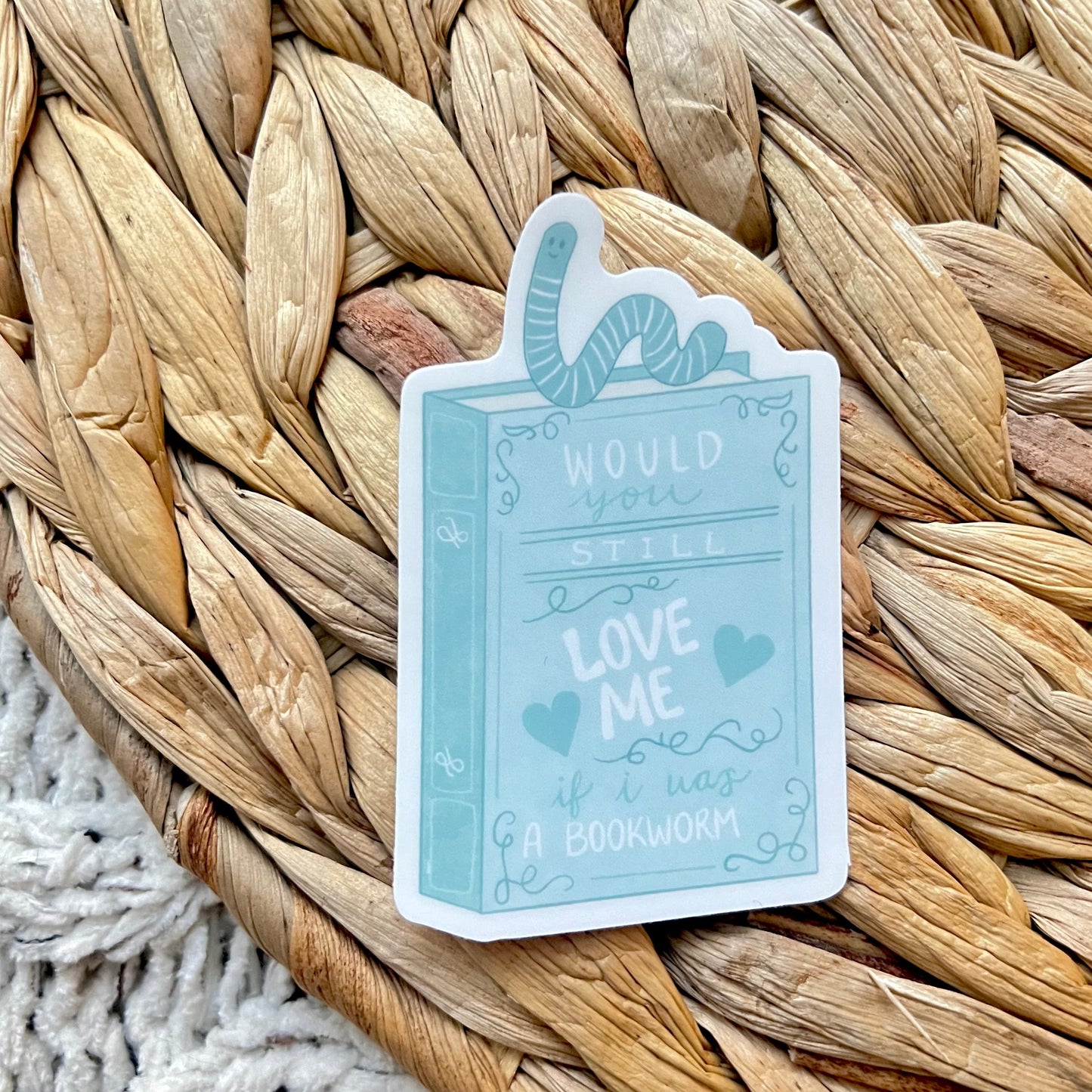 Sticker of a blue book with the words "Would you still love me if I was a bookworm?" on it and a worm on top of the book sitting on a woven mat