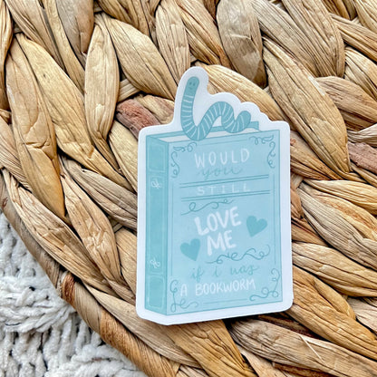 Sticker of a blue book with the words "Would you still love me if I was a bookworm?" on it and a worm on top of the book sitting on a woven mat