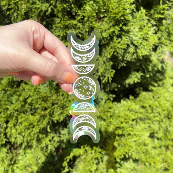 Hand holding up holographic suncatcher sticker shaped like the phases of the moon in front of a green plant, and it is refracting the light making rainbows