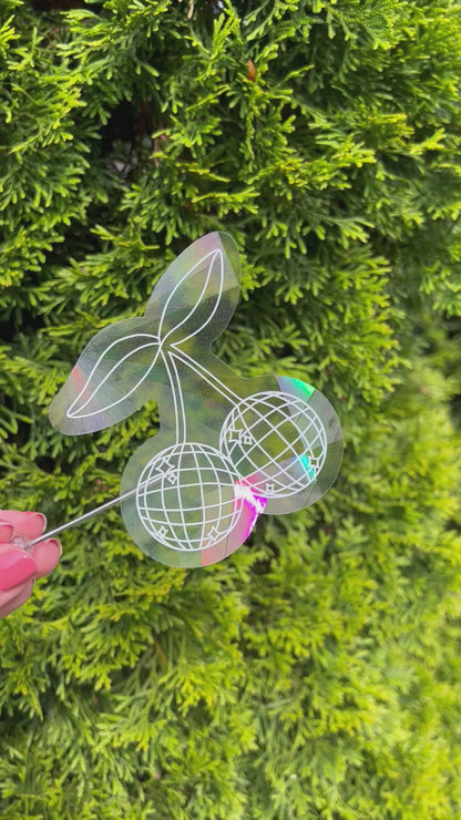 Disco cherry holographic suncatcher sticker held up refracting light and making rainbows in front of leafy green plant