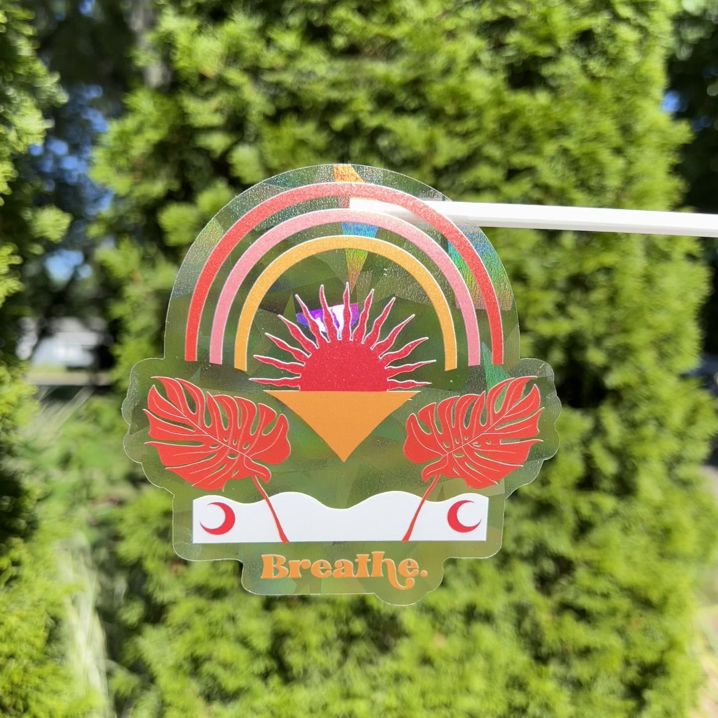Text saying "Breathe" with modern bohemian leafy and rainbow design on a holographic suncatcher sticker refracting light and creating rainbows in the sunshine
