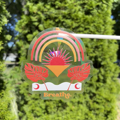 Text saying "Breathe" with modern bohemian leafy and rainbow design on a holographic suncatcher sticker refracting light and creating rainbows in the sunshine