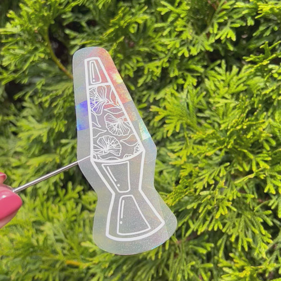 Holographic lava lamp suncatcher sticker held up refracting light and making rainbows in front of green foliage