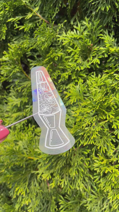 Holographic lava lamp suncatcher sticker held up refracting light and making rainbows in front of green foliage